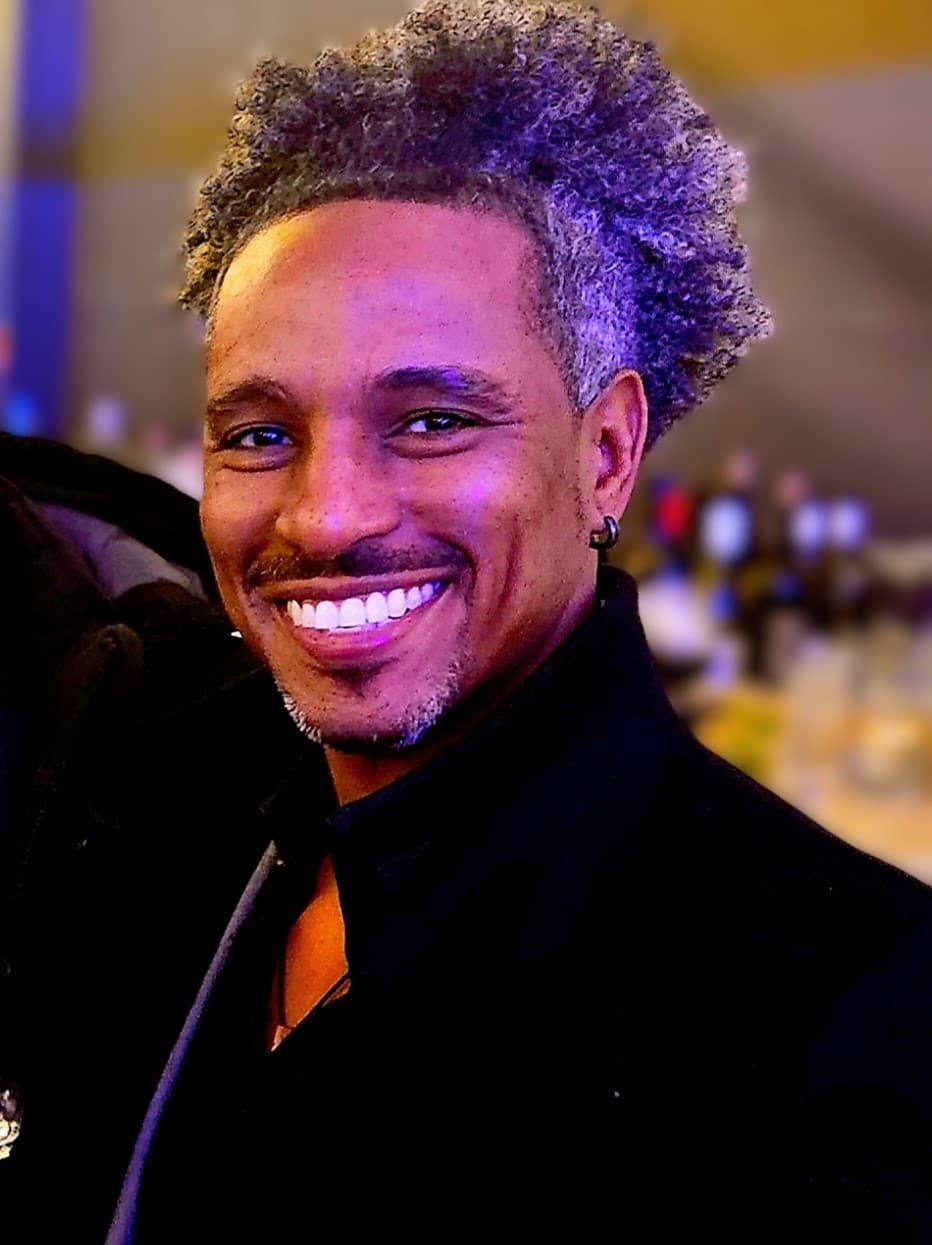 Smiling man with salt-and-pepper afro hair, mustache, and goatee, wearing a black jacket.