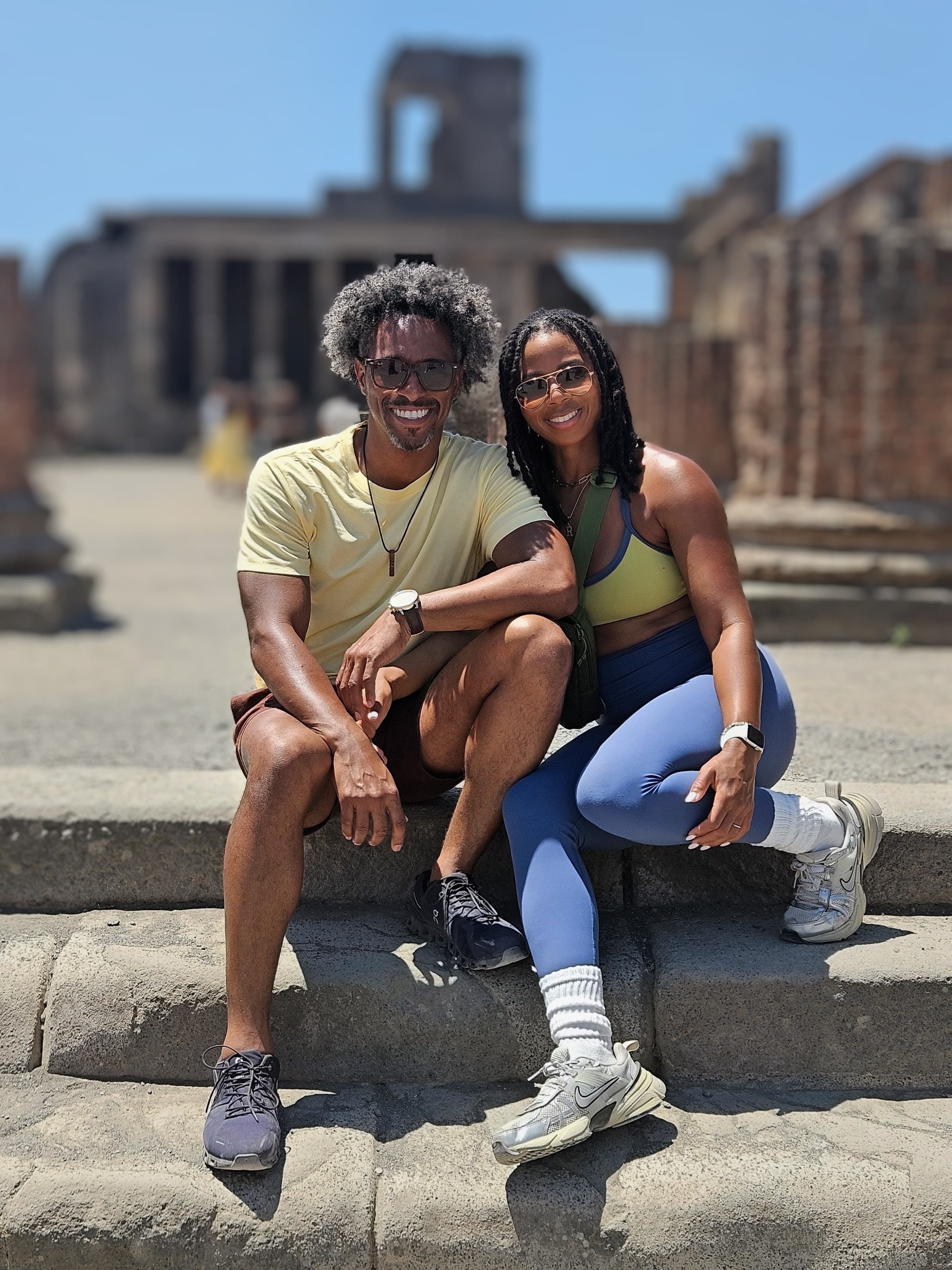 Smiling Black couple sits on stone steps in front of blurred ancient architectural ruins.