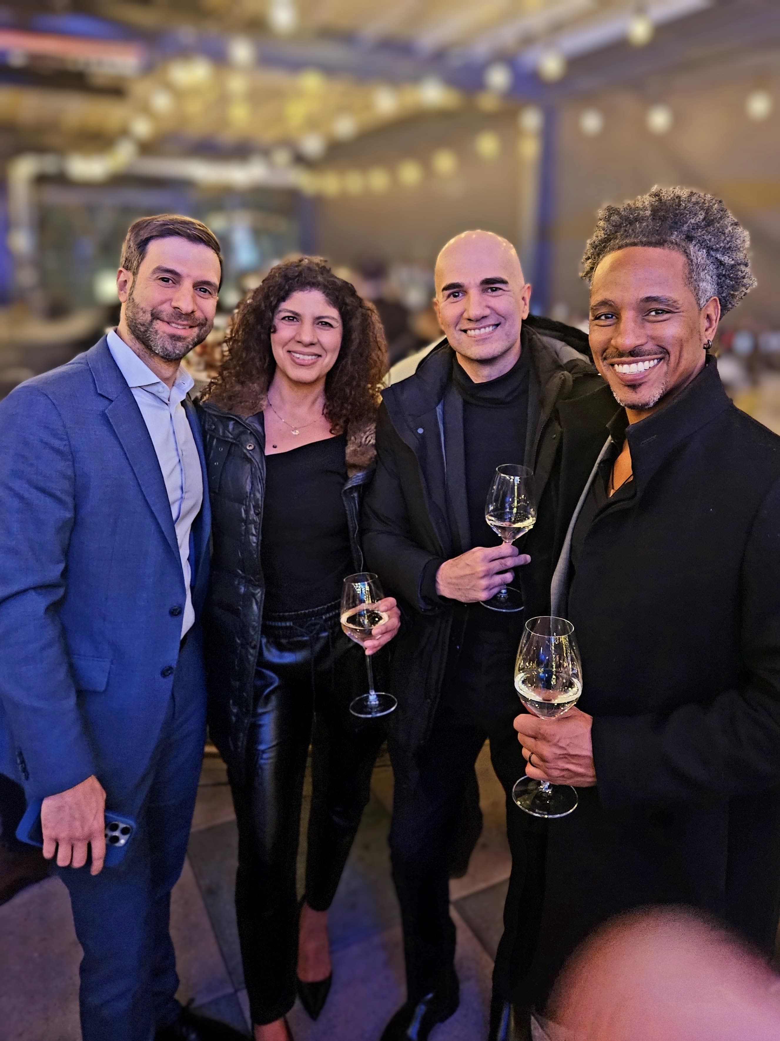 Four smiling people holding wine glasses at an outdoor evening event with blurred string lights.