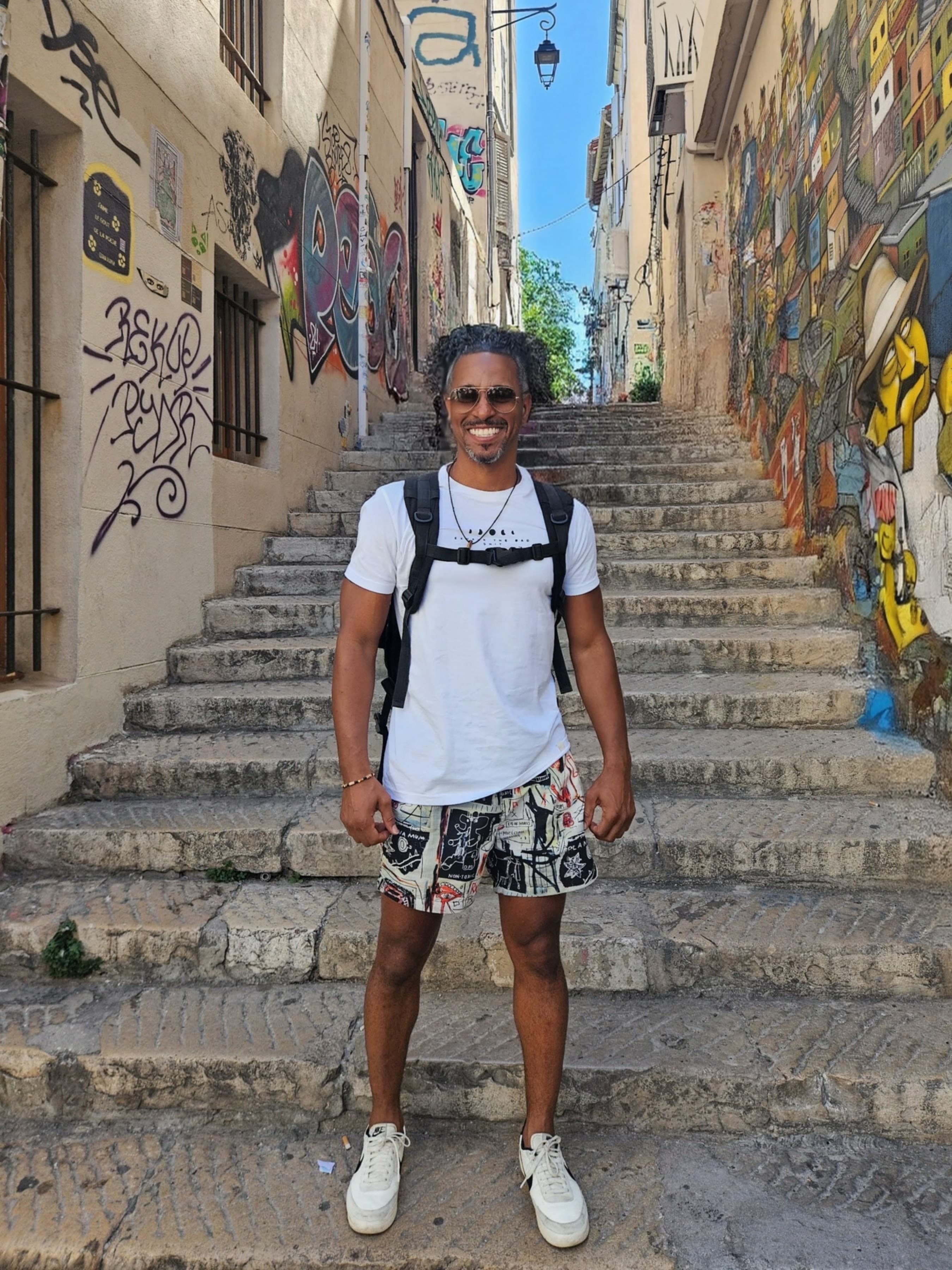Smiling man with a backpack stands on stone steps in a narrow, graffiti-lined alleyway.