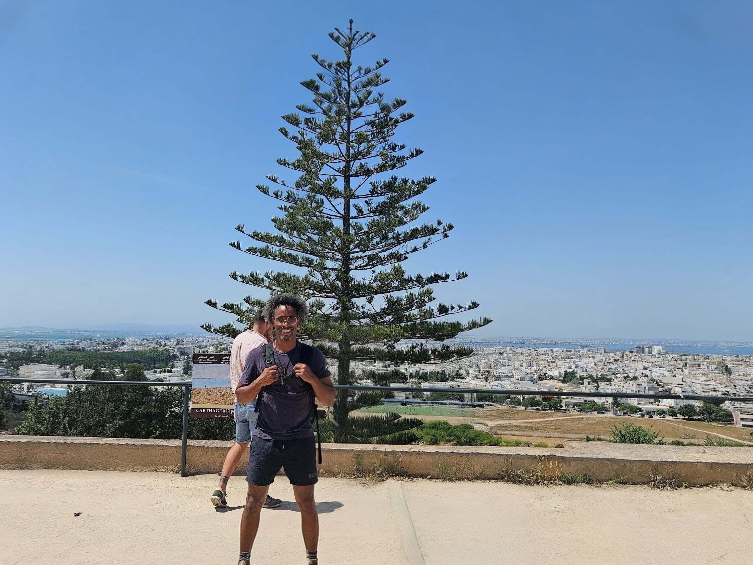 Smiling man with a backpack stands before a tall evergreen tree overlooking a coastal city.